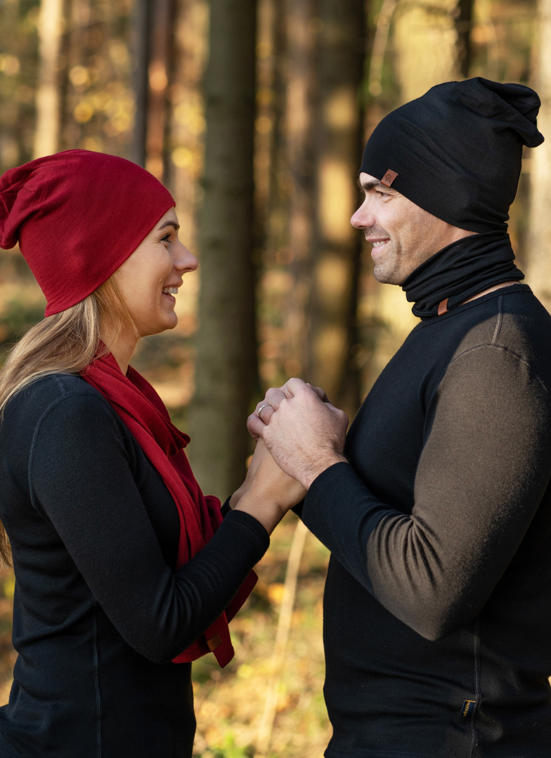 A man and woman stand face-to-face in a sunlit forest, holding hands. The woman wears a red beanie and scarf with a black top, while the man wears a black beanie and a dark puffer vest.