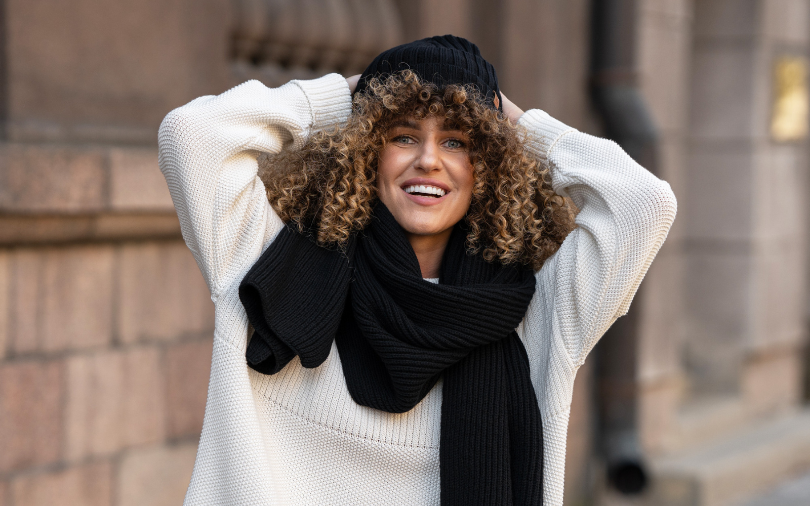 A woman with curly hair wearing a black set of knitted ribbed merino wool beanie and scarf.