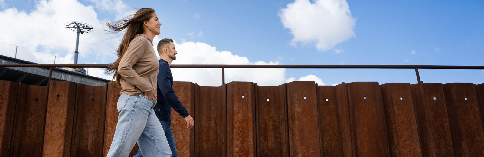 A smiling woman with long brown hair blowing in the wind walks outdoors alongside a man. She is wearing a tan hoodie and light blue jeans, while he is wearing a dark blue long-sleeved shirt. They are walking past a rusted corrugated metal wall under a bright blue sky with scattered white clouds.