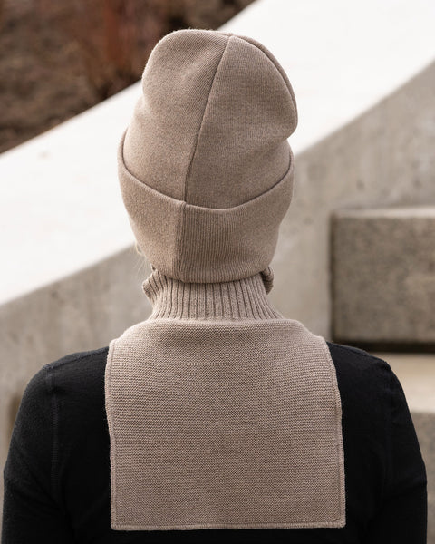 This image shows the back of a person's head and shoulders. They are wearing the same light brown or taupe knit beanie and neck warmer seen in the first image. The neck warmer's flat-knit panel is visible extending down the upper back over a black long-sleeved shirt. The background is slightly out of focus, showing a light-colored concrete structure, possibly part of the stairs seen in the other images.