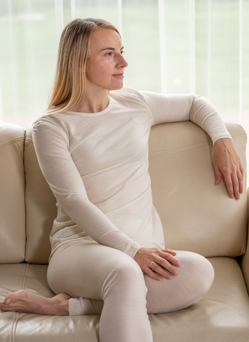 A vertical portrait of a blonde woman sitting on a light-colored sofa, wearing a cream-colored ribbed long-sleeved top and matching leggings, looking thoughtfully out of a window.