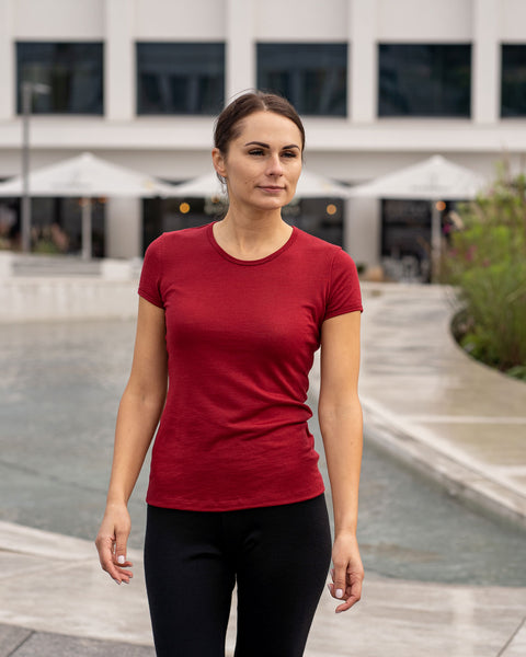 Woman wearing a merino wool menique royal cherry t-shirt and black pants standing in an outdoor setting with buildings and umbrellas in the background.