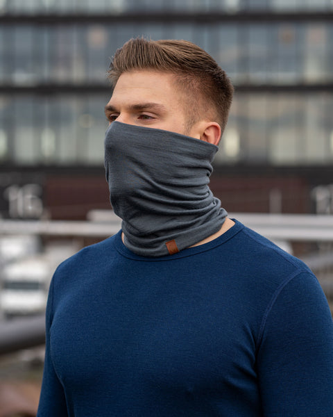 A man wearing the menique unisex neck gaiter in perfect gray color. He is outside, dressed in a long sleeve merino top in dark blue color.