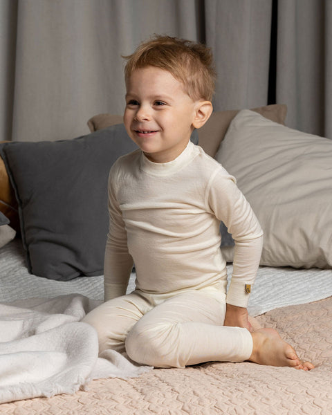 a boy sitting on a bed, wearing the Kids&#39; Merino 160 Long Sleeve Set in Natural color.