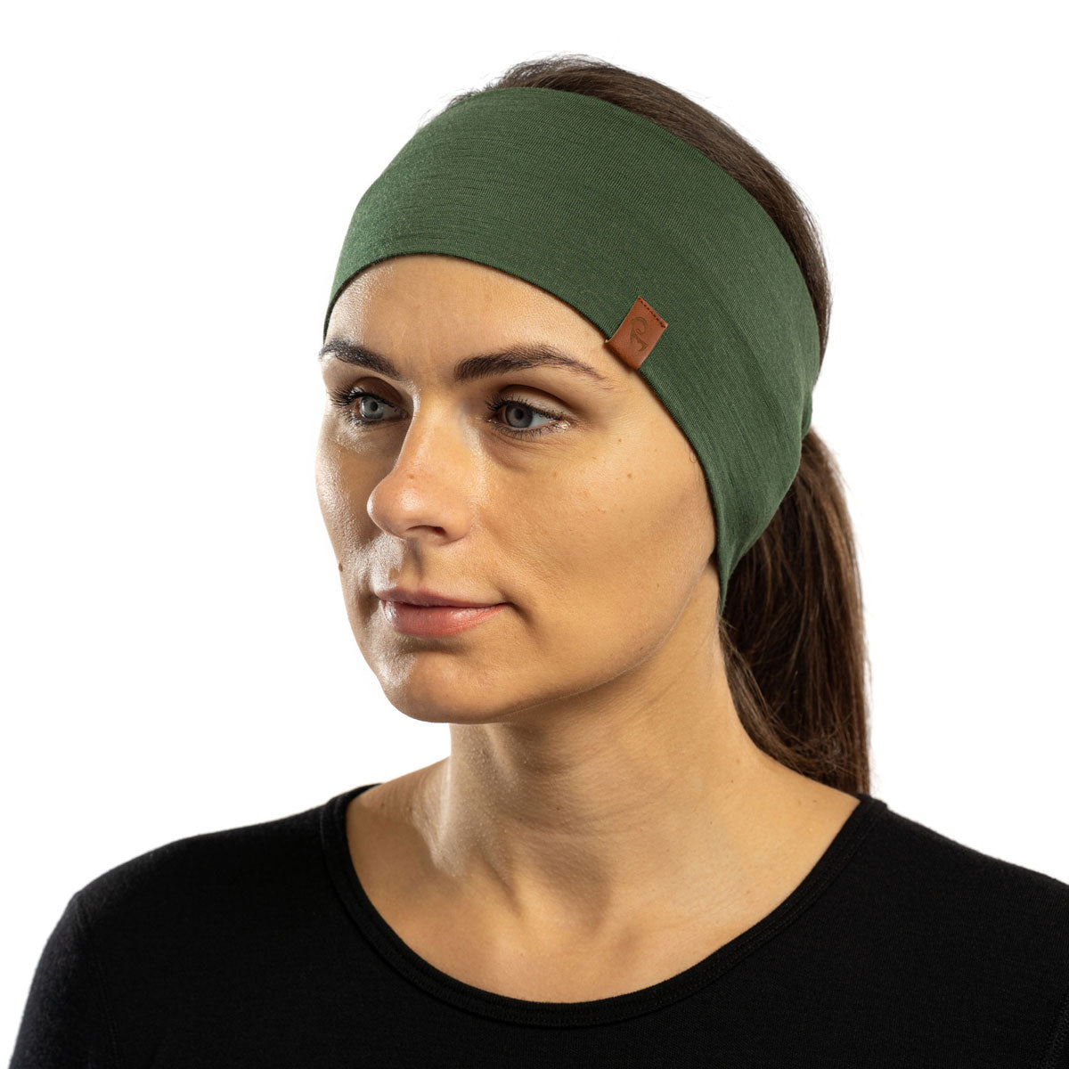 The picture features a woman sitting down in a forest, wearing the unisex dark green menique merino HEADBAND. She is also wearing a sleeveless black merino top. She is looking to the side.